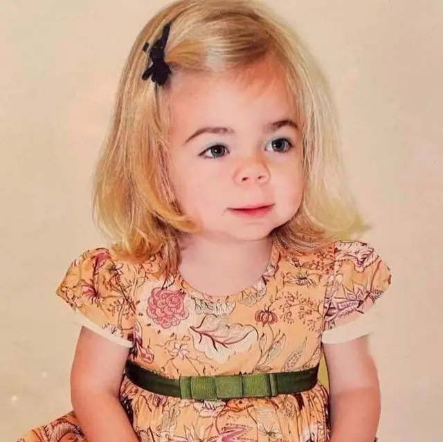 Sophia cuando era muy pequeña. Tiene el cabello rubio, a la altura de los hombros, sujeto con un lazo negro que lo aparta de su rostro. Mira fuera de cámara y lleva puesto un vestido marrón de tonos otoñales, de manga corta, con un lazo verde sobre una cinta que lo rodea.