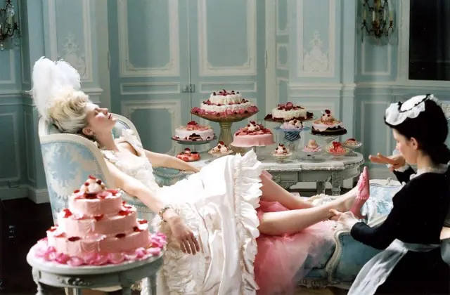 Fotograma de la película sobre María Antonieta, protagonizada por Kirsten Dunst, quien puede verse completamente reclinada en una silla, rodeada de pasteles, mientras una sirvienta le quita los zapatos.