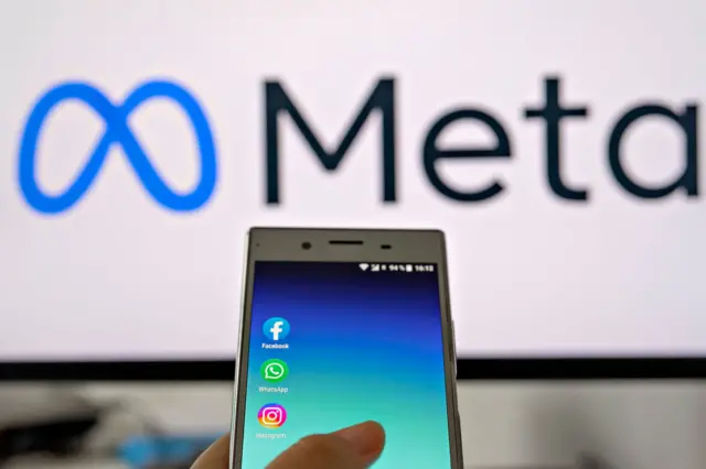 Una mano sujeta un teléfono con los íconos de las principales aplicaciones de Meta en la pantalla. El teléfono apunta al logo de la compañía, que se ve de fondo.
