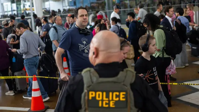 Agente de ICE frente a una fila de personas en un aeropuerto