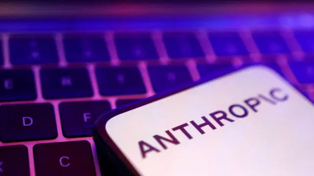 Una pantalla de smartphone que muestra el logotipo de Anthropic en letras negras sobre un fondo completamente blanco, colocada sobre un teclado de portátil iluminado en rosa y morado.