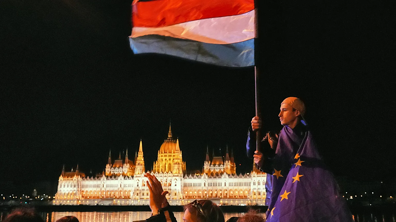  A man wrapped in the European Union flag waves a Hungarian flag, backdropped by the parliament building, early Monday April 13, 2026 as people celebrate Peter Magyar ousting 