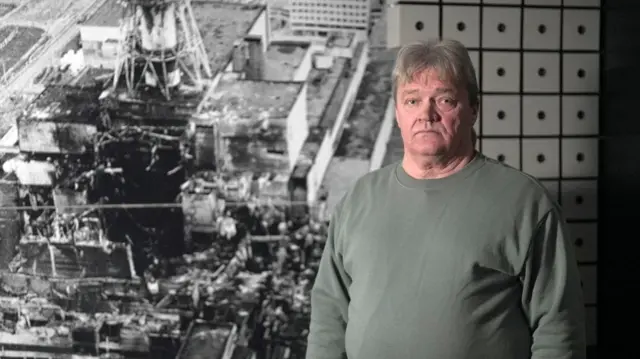 Nikolai Solovyov, ingeniero jefe de la sala de turbinas en el momento del accidente, fotografiado frente a una imagen del reactor destruido, en el museo de Slavutych. Tiene el pelo canoso, lleva una sudadera de color gris verdoso y mira a la cámara con expresión seria.