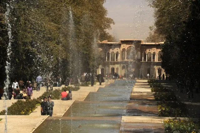 Un jardín rodeado de árboles con un canal de agua en el centro de donde salen fuentes. Hay personas sentadas, paradas y otras caminando al fondo se ve una estructura