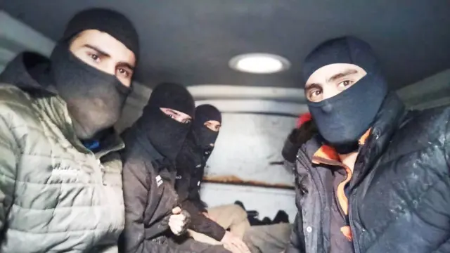 Un grupo de hombres con pasamontañas negros se sienta muy juntos en la parte trasera de una furgoneta con poca luz. Todos llevan una gruesa chaqueta de invierno y miran fijamente a la lente de la cámara.
