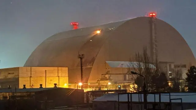 El gran escudo metálico curvado que cubre el reactor cuatro, fotografiado con poca luz, con llamas saliendo de un agujero en uno de sus lados, donde fue alcanzado por un dron el 14 de febrero de 2025. Se observan luces rojas en la parte superior de la cúpula y luces amarillas alrededor de los edificios más bajos situados cerca de ella.