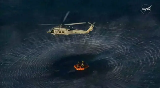 Un helicóptero de la marina sobrevuela una balsa que flota en el mar ycontiene a los tripulantes del Artemis II. La fuerza de sus aspas crea ondas concéntricas en el agua