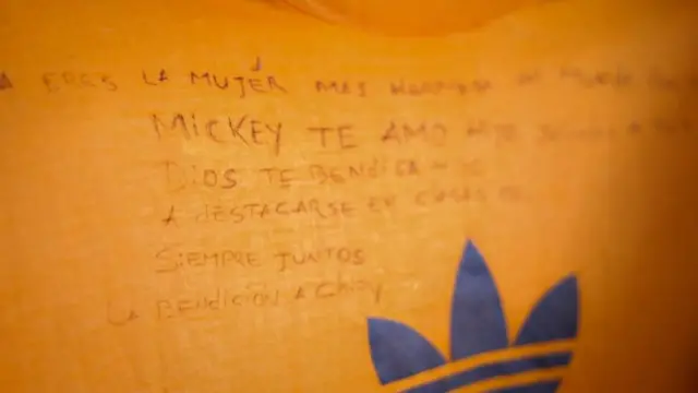 Una toma cercana del mensaje escrito con bolígrafo por Ángel Godoy en una camiseta. El mensaje dice a su esposa: "Eres la mujer más hermosa del mundo. Orgulloso de ti". También hay un mensaje para su hijo: "Te amo hijo. Saludos a tus amigos. Dios te bendiga hijo. A destacarse en clases. Siempre juntos".