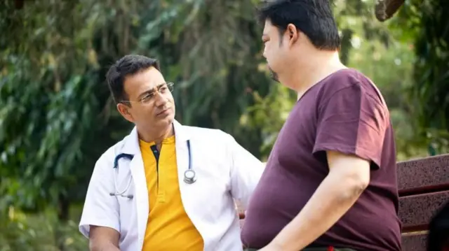Un hombre que viste una bata blanca y un estetoscopio mira a otro que tiene una barriga prominente.