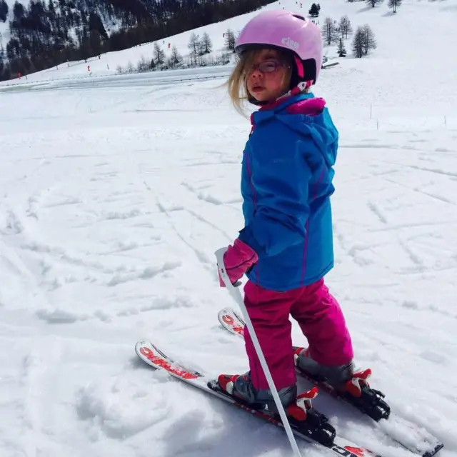 Sophia de niña, sobre esquís en la nieve. Tiene el cabello largo y rubio, lleva gafas y viste un casco rosa, una chaqueta impermeable azul y pantalones de esquí rosas. Sostiene un bastón de esquí y en sus esquís hay inscripciones en rojo. Está mirando a la cámara.