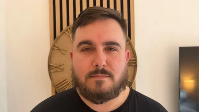 Primer plano de un joven, de medio cuerpo, con barba y bigote tupidos, vistiendo una camiseta negra. Tiene una expresión seria y hay un reloj de madera justo detrás de su cabeza.