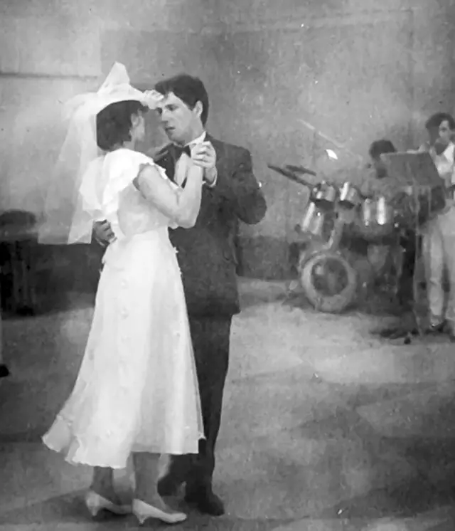 Iryna y Serhiy bailan el día de su boda. Ella lleva un sombrero blanco, un vestido blanco vaporoso y zapatos blancos sin cordones de tacón bajo. Él lleva un traje oscuro. La mira a los ojos, pero con expresión inquieta.