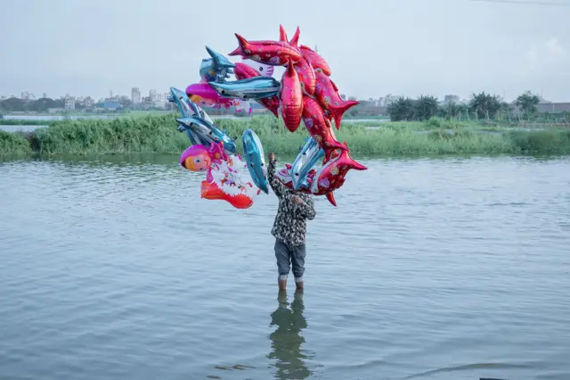 Un hombre de pie en un río, sosteniendo globos de colores vivos por encima de su cabeza.