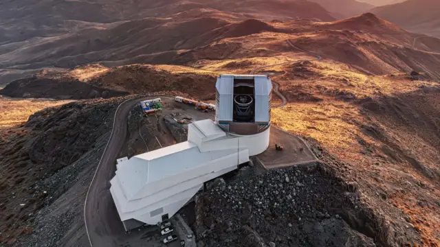 Vista aérea de un enorme telescopio a través de su vitrina, situado en lo alto de un gran edificio blanco al borde de una cima rocosa. Un camino rodea el observatorio y algunos coches se ven en la parte inferior, mientras el paisaje montañoso se tiñe de un tono dorado al atardecer.