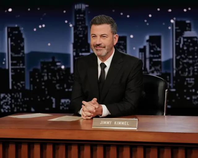 Hombre sentado detrás de un escritorio de programa nocturno, con traje oscuro y manos entrelazadas; fondo de ciudad nocturna y rótulo “JIMMY KIMMEL” en el escritorio.