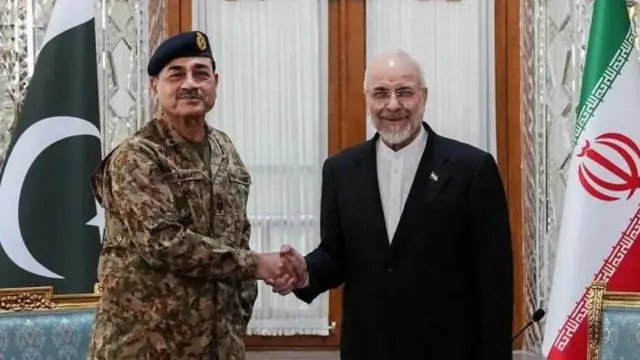 El presidente del Parlamento iraní, con una chaqueta oscura sobre una camisa blanca, estrecha la mano del jefe del Ejército de Pakistán, que viste uniforme militar y una boina verde. Ambos hombres sonríen, y las banderas de ambos países se ven a los lados de la imagen.