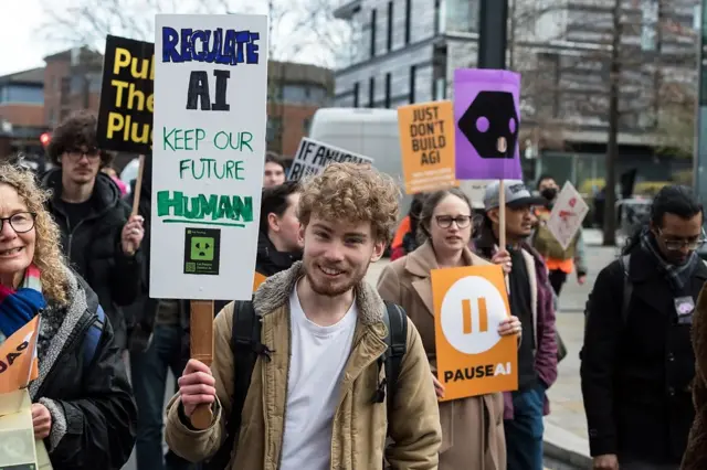 Un joven sonriente sostiene un cartel pidiendo que se regule la IA, para mantener un "futuro humano", en medio de una protesta.