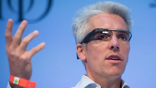 Un hombre en el escenario lleva puesto el intento inicial de Google de gafas inteligentes, las cuales presentan un cable prominente a través de la frente y una pequeña cámara frente a uno de los ojos.
