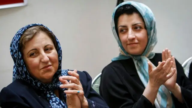 Shirin Ebadi y Narges Mohammadi aplauden durante una conferencia de prensa. Ebadi lleva un pañuelo estampado en azul oscuro, con algo de cabello visible, mientras que Mohammadi lleva un pañuelo estampado en azul claro, con parte de su cabello visible, y una chaqueta negra.