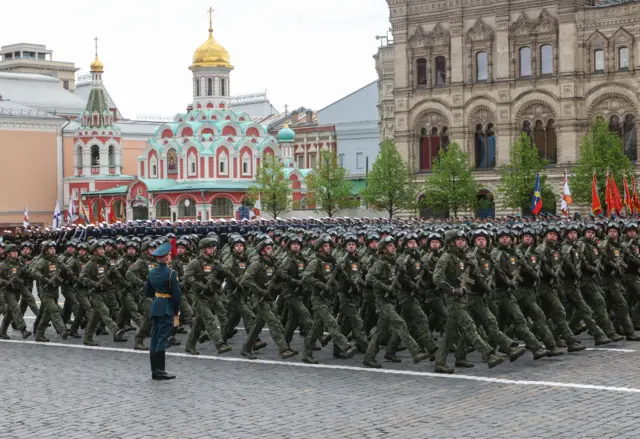 Unos militares rusos desfilan en filas vestidos con uniformes de camuflaje verde oscuro, cascos con gafas protectoras colocadas encima y empuñando armas. Detrás de ellos se divisa la catedral de Kazán, pintada en tonos verde azulado, rojo y blanco, y coronada por una cúpula dorada.