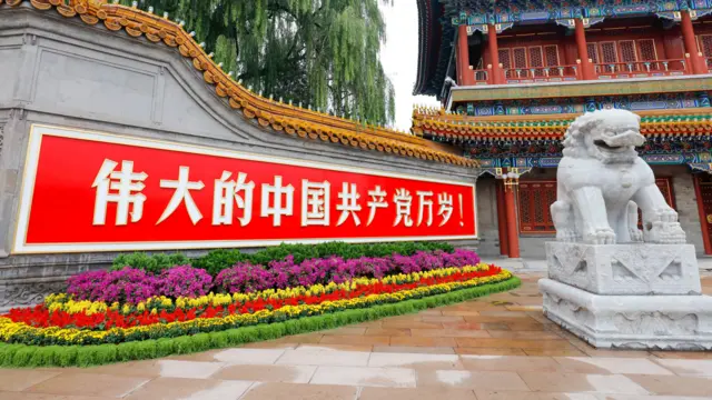 Un largo letrero rojo con caracteres chinos blancos cuelga de un muro de piedra, con un colorido parterre debajo; un león de piedra blanca descansa sobre un pedestal a su lado, con un edificio ornamentado visible al fondo.