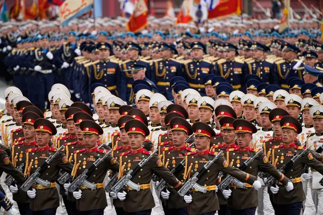 Soldados norcoreanos desfilan en filas portando armas. Los soldados de las primeras filas visten uniformes de color caqui con galones dorados y rojos. Más atrás se encuentran otros soldados norcoreanos con uniformes blancos con galones dorados y negros. Detrás de ellos hay filas de soldados rusos.