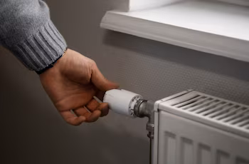 Primer plano de una mano girando el regulador blanco de un radiador metálico de color claro, situado junto a una pared gris y un alféizar blanco