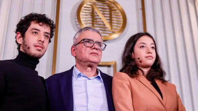 Taghi Rahmani, Ali Rahmani y Kiana Rahmani posan para una fotografía durante una conferencia de prensa en el Instituto Nobel. El logotipo dorado del Nobel se ve en el fondo. Los tres llevan pequeños tocados con micrófono.