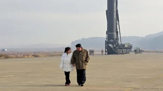 Kim Ju Ae con cabello largo y una chaqueta blanca camina tomada de la mano junto a su padre: Kim Jong Un. Detrás se ve una plataforma para lanzar un misil balístico. 