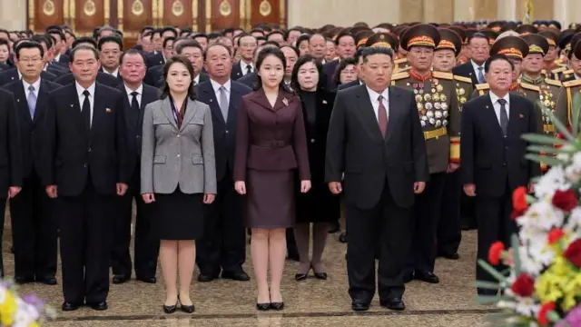 Ri Sol Ju, vestida con un traje de chaqueta gris y falda, junto a su hija, quien viste un traje similar morado y al lado el líder coreano durante un homenaje al fundador del país.