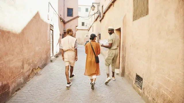 Dos hombres van caminando junto a una mujer por lo que pareciera ser un destino turístico en Medio Oriente. El hombre de la derecha lleva un sombrero y un mapa en las manos.