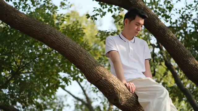 Un hombre vestido de blanco y sentado en la rama de un árbol, mira pensativamente hacia el suelo.