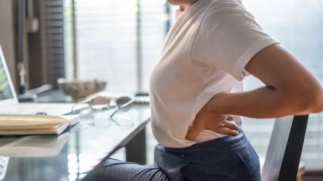Una mujer sentada en un escritorio se lleva las manos a la espalda en señal de dolor