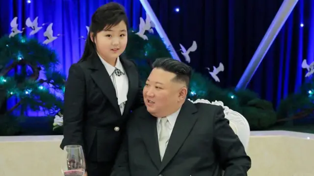 Kim Ju Ae, de pie junto a su padre, Kim Jong Un, ambos aparecen vistiendo trajes negros con camisa blanca.