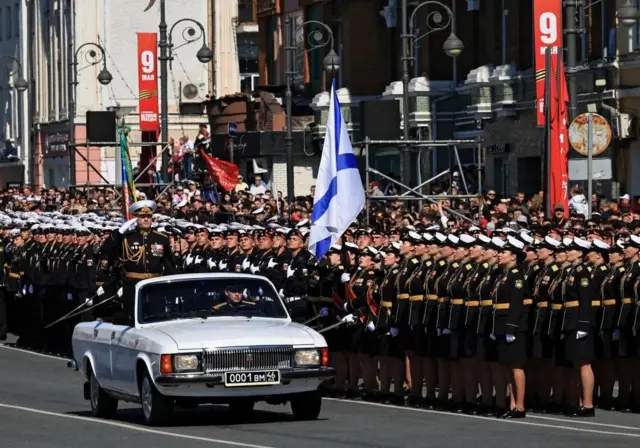 Un comandante militar ruso saluda mientras se encuentra de pie en un coche blanco descapotable conducido por otro soldado, en una carretera flanqueada por filas de militares.