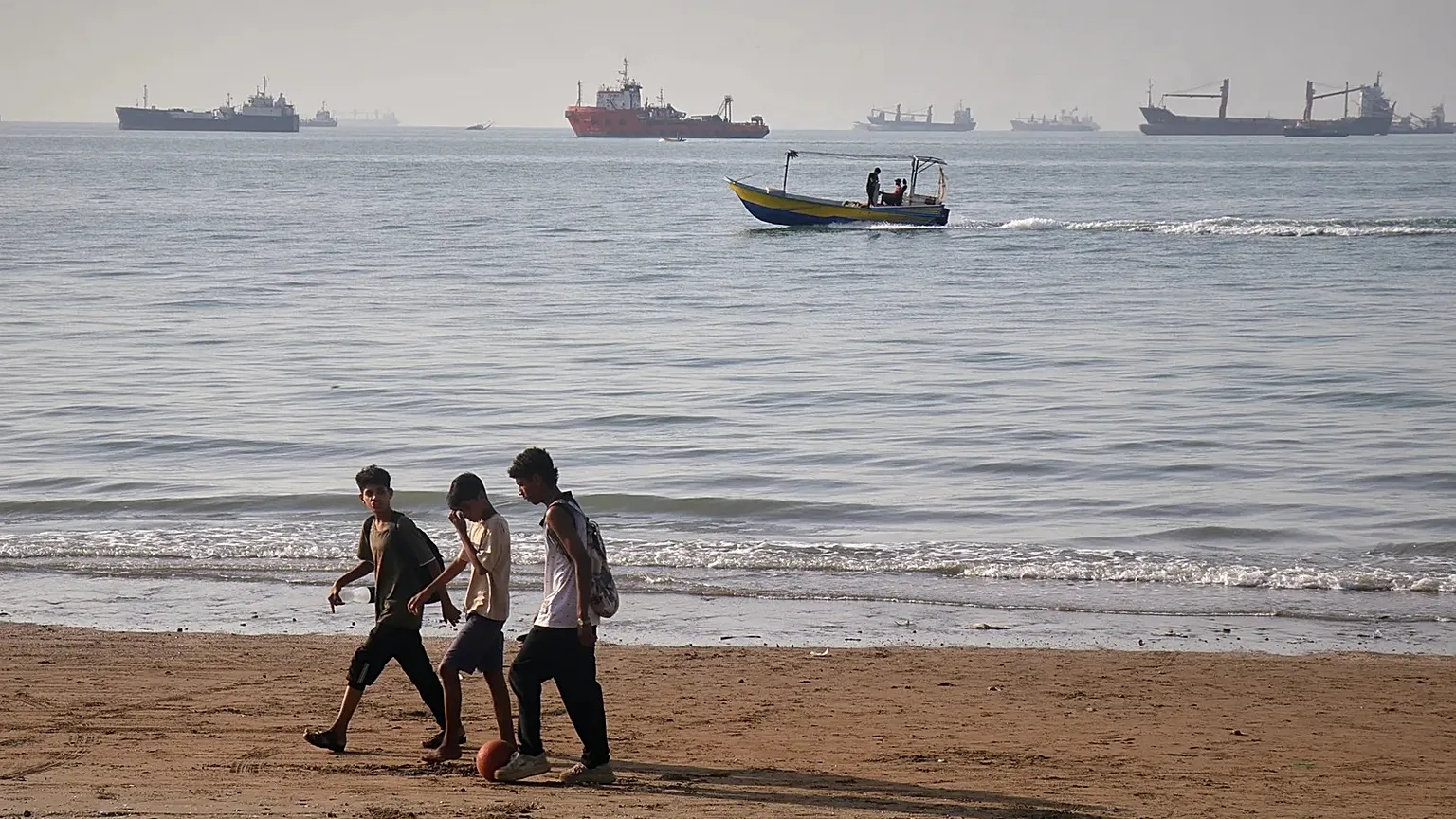 Children walk along the shore as a motorboat passes offshore and bulk carriers in the Strait of Hormuz, 27 April, 2026