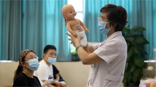 Una médica con una mascarilla sostiene un muñeco de bebé durante una clase para padres de recién nacidos mientras una mujer y un hombre observan.