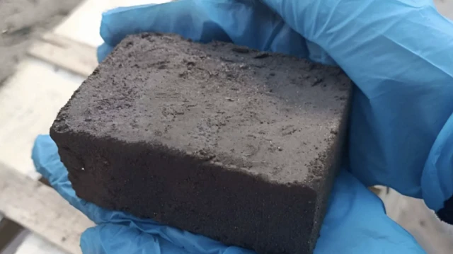 Una foto cerrada de un gran bloque de color gris oscuro, sostenido por un par de manos con guantes de goma azules.
