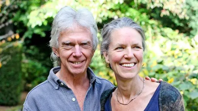 Foto de una mujer y su marido sonriendo. El marido tiene la mano sobre el hombro de su mujer. Los dos van vestidos de azul. Ambos tienen el pelo canoso. Detrás de ellos se ven árboles y hojas.