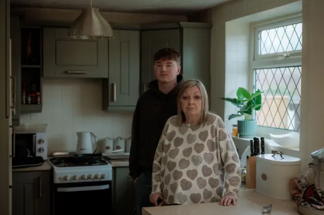 En primer plano, Susan, la abuela de Alex está de pie en la cocina y detrás de ella está Alex.