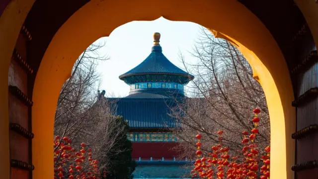 Una vista a través de un portal histórico, contemplando los ornamentados edificios de un templo en tonos azules, rojos y dorados, con numerosas linternas chinas colgando de los árboles desnudos en primer plano.