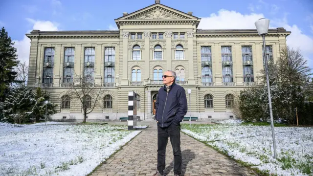 Un hombre está de pie frente a un edificio con nieve en el césped.