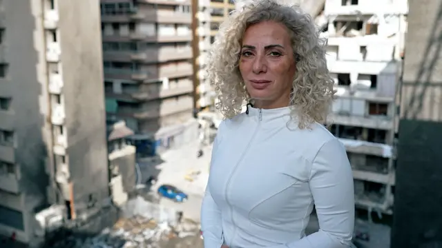 Una mujer parada en un balcón, con moretones en la cara, desde donde se mira la destrucción causada por una bomba.