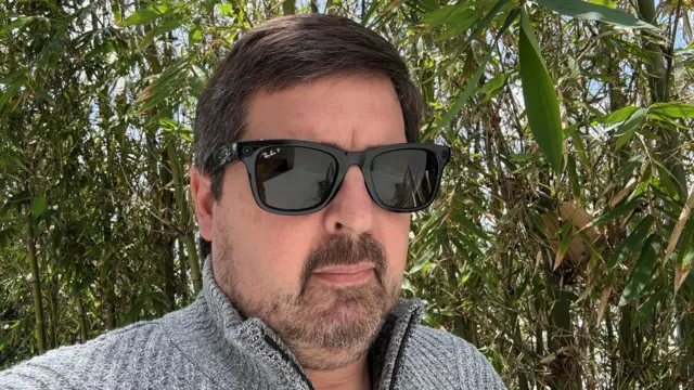 Un hombre de pie al aire libre, con un suéter gris y unas gafas Ray-Ban de Meta, con una pared de bambú verde detrás de él.