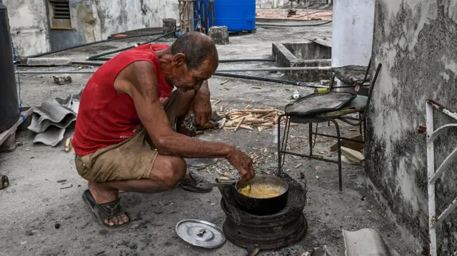 Un hombre, con camisa rosa y un pantalón corto beige, está agachado cocinando en un a olla negra con leña durante un apagón en La Habana