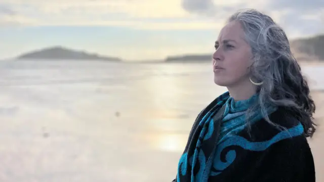 Una mujer de cabello gris y negro contempla el mar. Lleva un llamativo pañuelo con motivos azules y negros. La orilla aparece desenfocada a sus espaldas mientras brilla el sol.