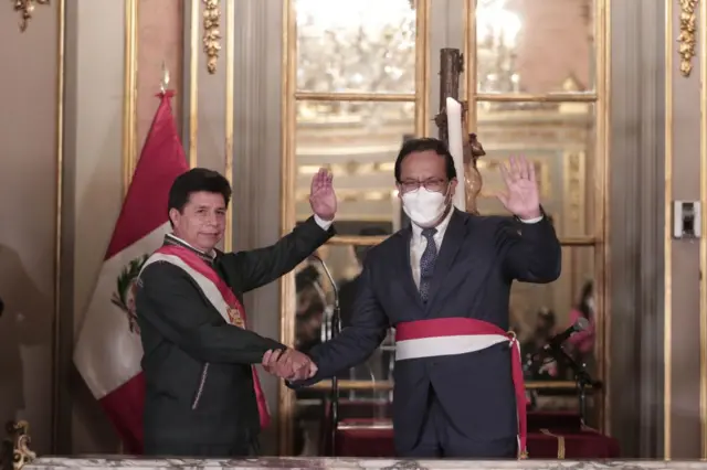 Pedro Castillo y Roberto Sánchez (d) saludan sonrientes con las manos de ambos estrechadas. Ambos lucen bandas con los colores de la bandera peruana. Sánchez lleva una mascarilla sanitaria.