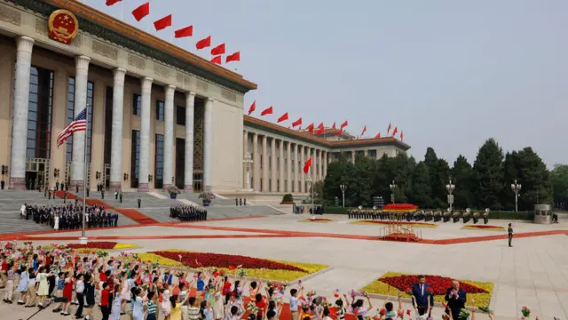 Una vista amplia de una plaza pavimentada con banderas chinas y estadounidenses izadas, y un largo edificio con columnas a la izquierda que luce banderas chinas en su tejado; en primer plano, se observa a los presidentes Xi y Trump saludando a la multitud.