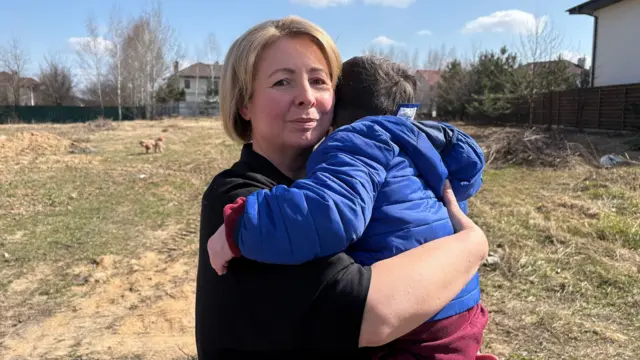 Una mujer sostiene a un niño pequeño en brazos. El niño está de espaldas a la cámara, por lo que no se ve su rostro. Tiene el pelo oscuro y lleva una chaqueta azul y pantalones rojos. La mujer mira a la cámara; tiene el pelo corto, liso y rubio, y lleva una blusa negra. Están al aire libre y se puede ver hierba alta y algunas casas detrás.