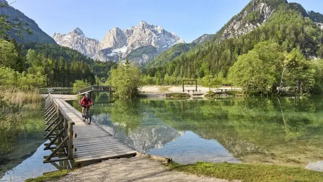 Una persona mayor con una chaqueta roja anda en bicicleta sobre un puente de madera que se extiende por un río en un hermoso parque natural